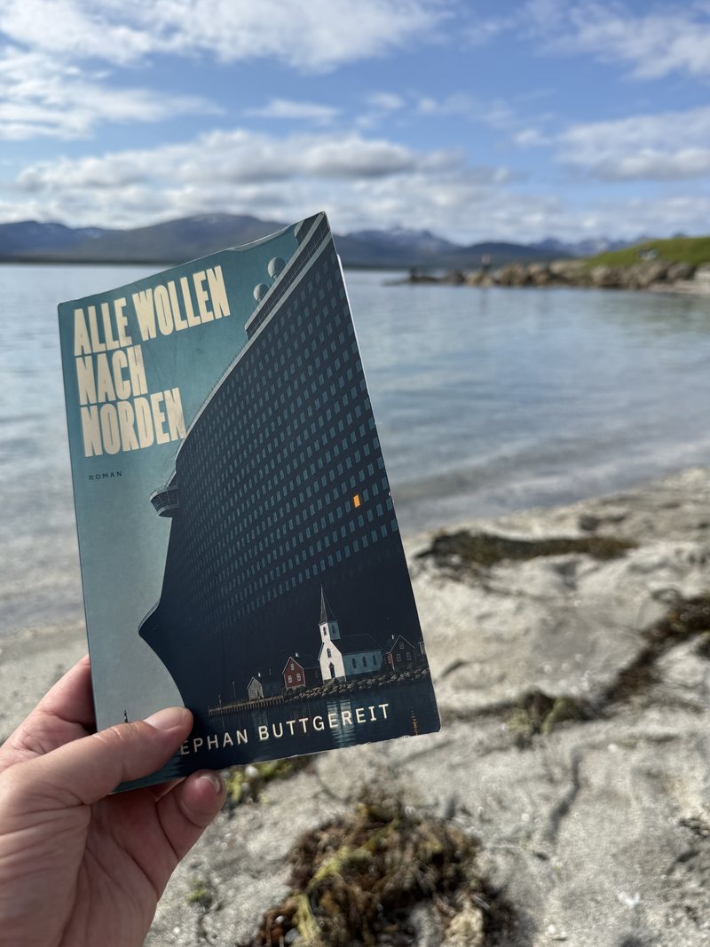 Das Taschenbuch Alle wollen nach Norden liegt auf Felsen am Stadtstrand Telegrafbukta in Tromsø, dahinter das Wasser des Sunds